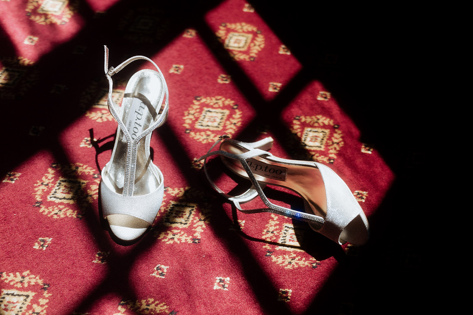 White satin open-toe bridal heels with crystal ankle straps on red patterned carpet with geometric shadow lines