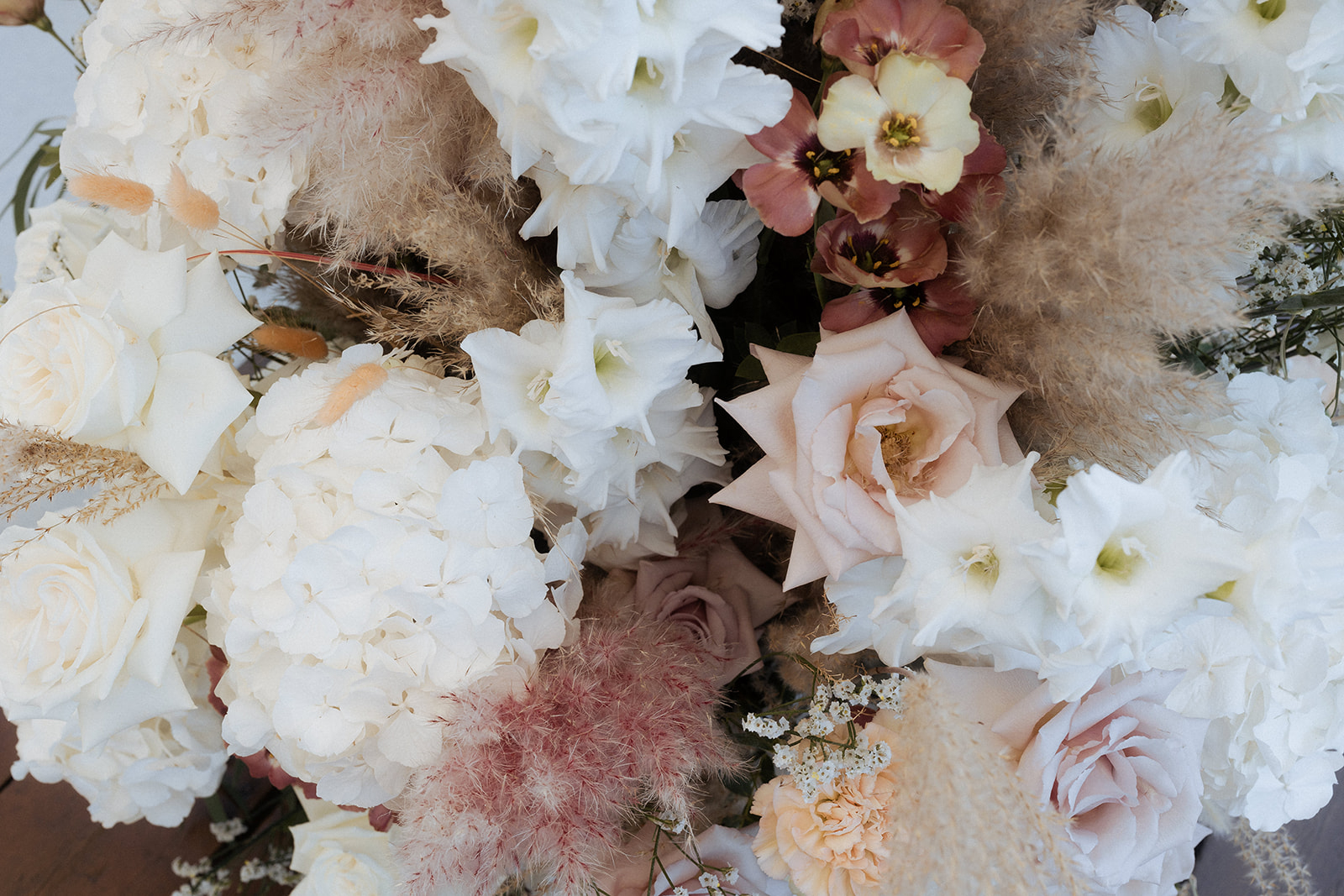 Close-up detail shot of a large wedding floral arrangement featuring a mix of white hydrangeas, white gladioli, cream and blush roses, mauve roses, dusty rose smoke bush (cotinus), pampas grass, burgundy-edged tulips, peach carnations, and white gypsophila. The overall color palette is soft and muted, combining ivory, blush, mauve, and warm beige tones in a boho-romantic style. No people are visible; the frame is entirely filled with the floral arrangement.
