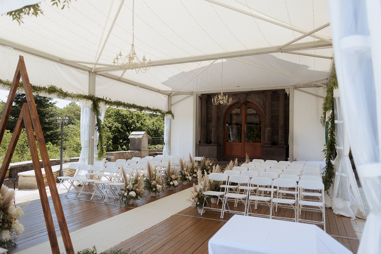 An empty outdoor ceremony setup photographed from a wide-angle elevated perspective, showing a white marquee tent erected on a wooden deck terrace against the facade of a château with classical stone columns and large arched wooden doors. Rows of white folding chairs are arranged on either side of a cream fabric aisle runner, with floral arrangements placed along the aisle featuring pampas grass, blush and white blooms, and dried botanicals in a boho-natural style. Two gold chandeliers hang from the white tent ceiling, and the marquee posts are draped with green foliage garlands and sheer white curtains. A triangular wooden arch is partially visible in the foreground left. The overall decor palette is ivory, white, and natural beige with greenery accents. Potential venue feature image.