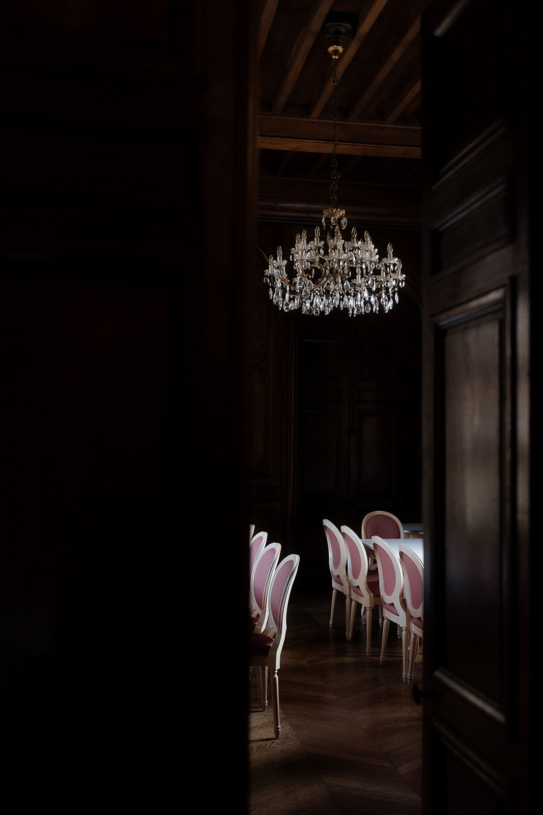 A moody interior shot of a formal dining room viewed through a partially open dark wooden door, with no people present. The room features a gold and crystal chandelier hanging from a dark wood-beamed ceiling, casting warm light in an otherwise deeply shadowed space. A long dining table is set with a white tablecloth and surrounded by Louis XVI-style medallion chairs with white painted frames and dusty rose pink upholstered seats and backs. The herringbone parquet floor and dark wood-paneled walls suggest a classic French château interior. Wide shot framed as a peek through the doorway, with strong contrast between the deep foreground shadow and the softly lit room beyond. Potential venue feature image.