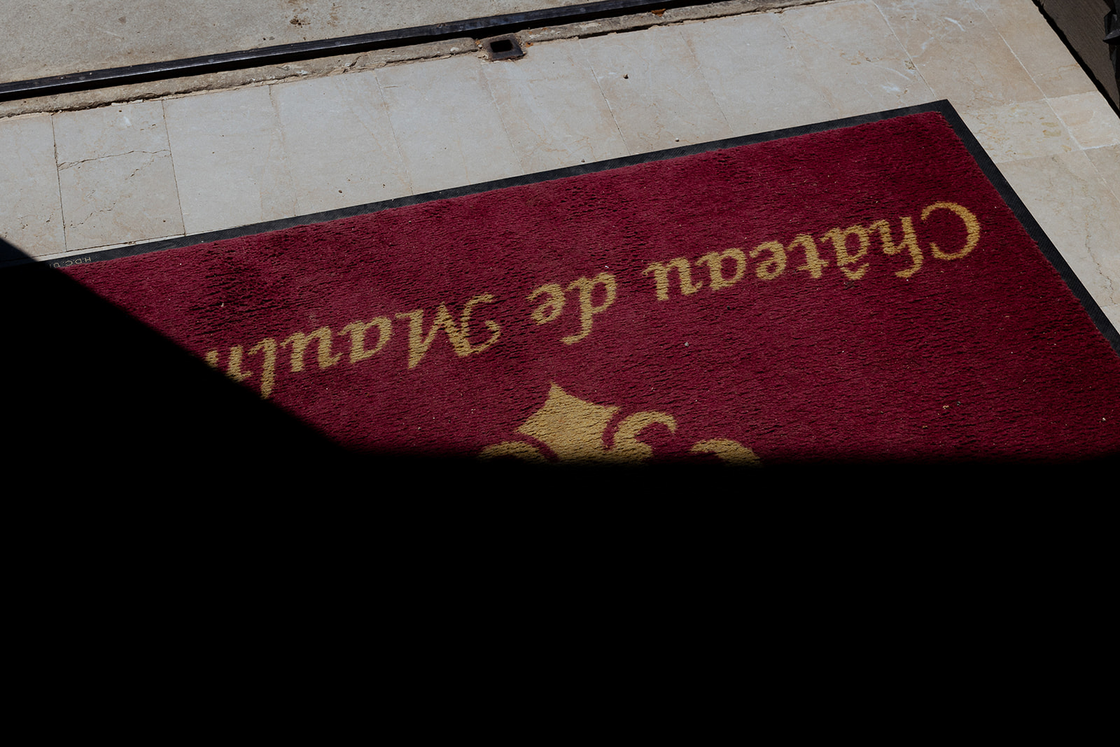 Close-up detail shot of a deep burgundy entrance mat with gold script lettering reading 'Château de Mauli...' (partially obscured) and a partial gold fleur-de-lis motif, positioned on cream-toned stone tile flooring. The image is shot at an angle with a strong diagonal shadow cutting across the lower half of the frame, creating high contrast between the lit and unlit portions of the mat. No people are visible. The shot serves as a venue identifier detail, typical of arrival or establishing imagery.