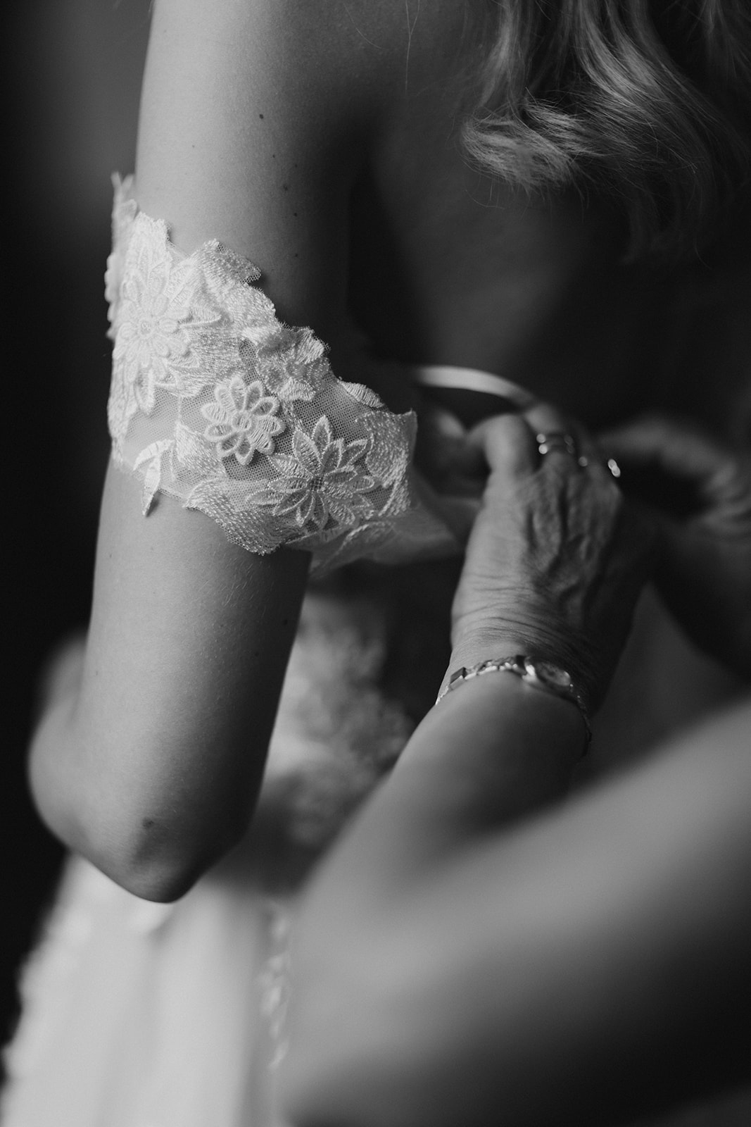 A black-and-white close-up detail shot captured during bridal preparations, showing an older person's hands — wearing a delicate bracelet and a ring — adjusting a lace garter on the bride's thigh. The garter features floral embroidery on a sheer tulle base. The bride has wavy blonde hair visible at the top of the frame, and the background is softly blurred with dark tones, drawing full focus to the garter and the hands. The high contrast and shallow depth of field emphasize the intergenerational moment of assistance.