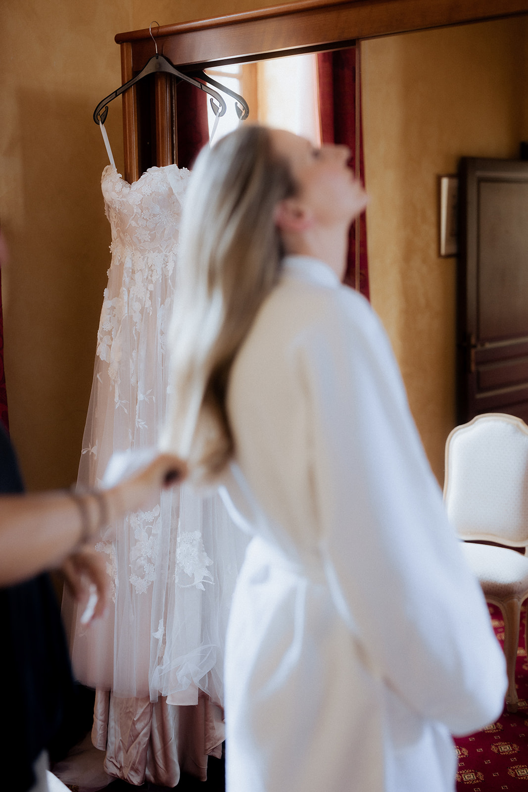 Bride in robe having hair styled with 3D floral lace applique gown hanging sharp in background focus