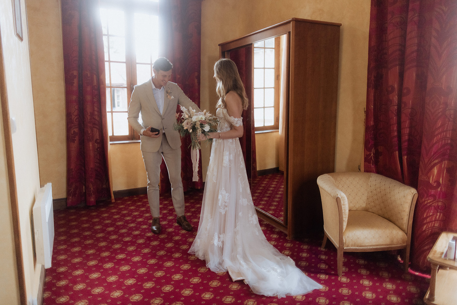 Groom in tan suit approaching bride holding garden-style bouquet during first look in chateau room