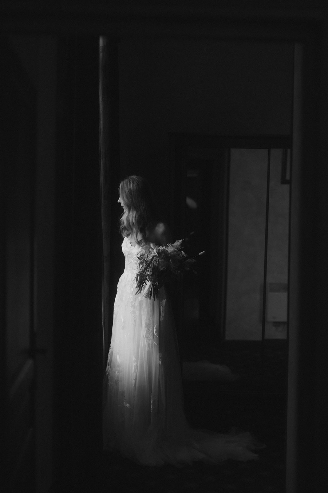 Black-and-white bridal portrait shot through a doorway with bride in lace gown holding a trailing bouquet