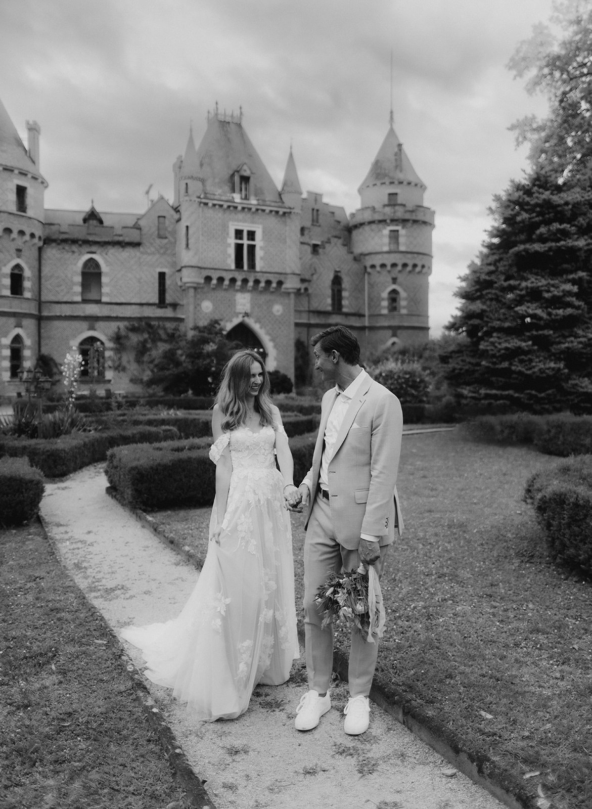 Black and white couple on gravel path before Gothic-turret chateau with lace applique gown