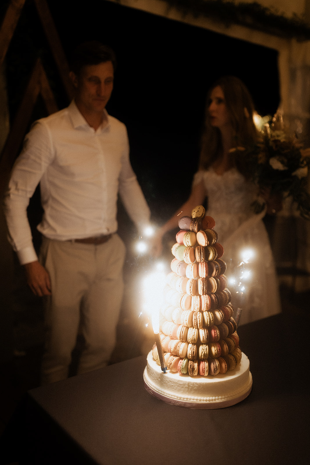 The couple stands behind a tall French macaron tower during an indoor evening reception, with the bride reaching toward the top of the tower. The macaron tower is the primary focus of the shot, stacked in a conical pyramid shape with macarons in pastel shades of mint green, dusty rose, ivory, and caramel, presented on a white cake board base with sparklers lit around it, creating bright bursts of light against the dark background. The groom wears a white dress shirt with light grey trousers, and the bride wears a white lace long-sleeve gown; a floral arrangement with greenery is partially visible to the right. The composition is a medium shot with shallow depth of field, keeping the macaron tower sharp in the foreground while the couple is softly blurred behind it, illuminated by warm, low-level reception lighting.
