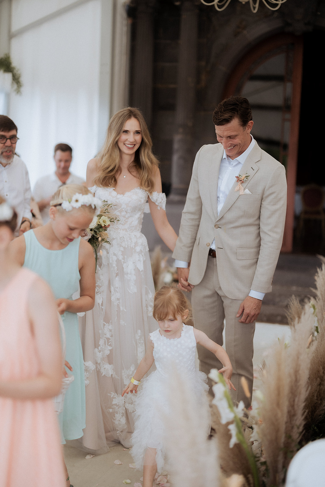 The bride and groom walk hand-in-hand down the aisle following the ceremony, surrounded by two young flower girls. The setting appears to be an outdoor or semi-covered terrace of a grand stone building with arched architectural details visible in the background. The bride wears an off-the-shoulder white gown with floral lace appliqué overlay on a nude base, and carries a loose bouquet of cream and blush flowers including what appear to be ranunculus and greenery; the groom wears a sand-colored linen suit with a light blue shirt and a small blush boutonniere. One flower girl wears a mint green dress with a white floral crown, while a younger flower girl wears a white textured tulle dress. The aisle is lined with arrangements of pampas grass, white blooms, and dried botanicals, contributing to a relaxed, boho-inspired aesthetic. The overall color palette is neutral and warm — ivory, sand, blush, and mint. Medium portrait-style shot with guests visible and slightly blurred in the background.
