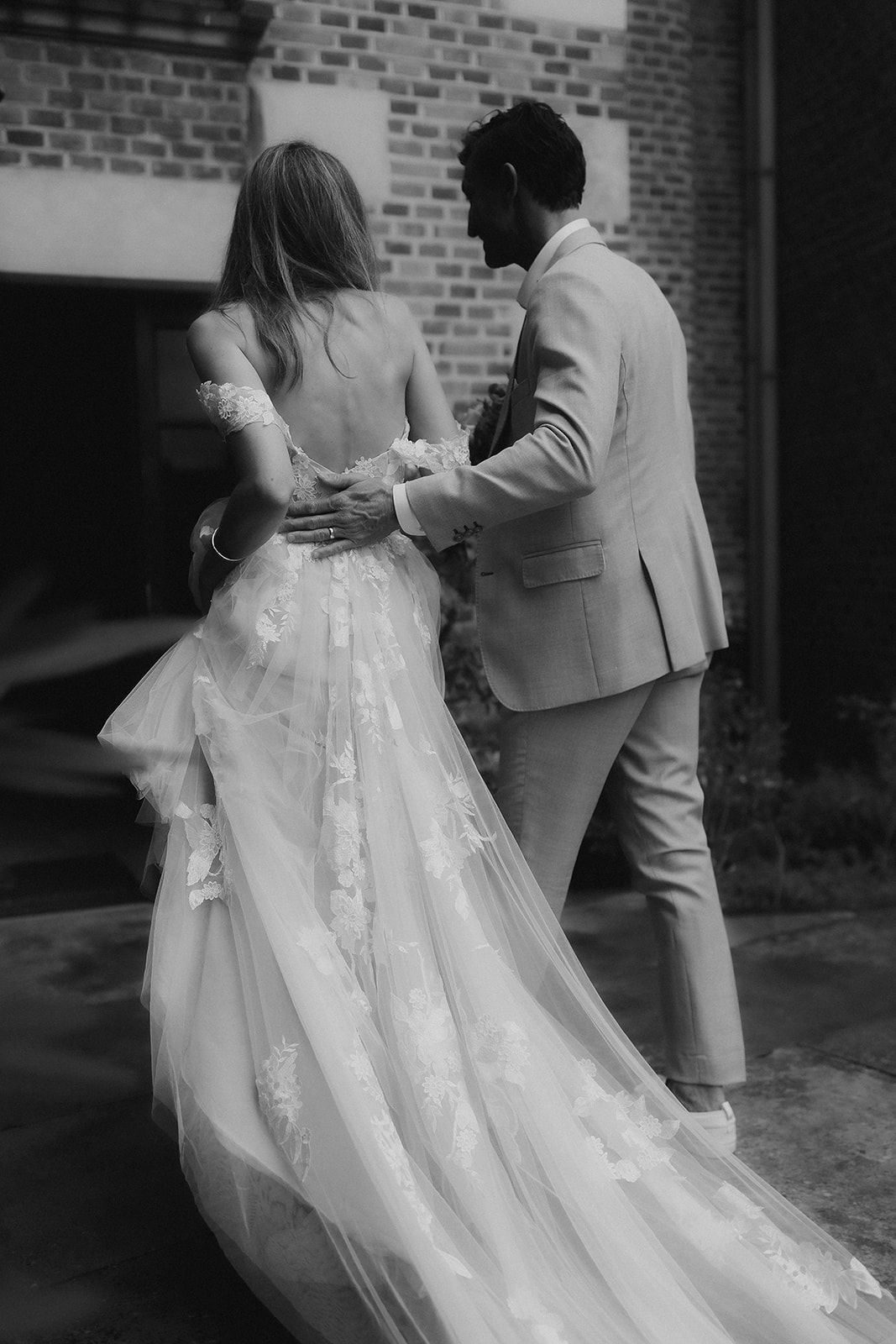 This is a black-and-white couple portrait shot from behind, showing the bride and groom walking together in front of a brick building exterior. The groom, wearing a light-toned slim-fit suit, has his hand placed on the bride's lower back. The bride wears an off-the-shoulder gown with a low open back, featuring floral appliqué lace detailing on layered tulle, with a full train spreading across the ground. The high-contrast black-and-white tones emphasize the texture of the lace appliqués and the volume of the tulle skirt. The composition is a medium portrait shot framed from ground level to capture the full length of the dress train.