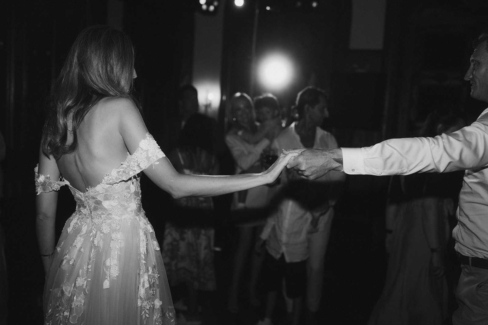 This black-and-white image captures the couple's first dance at an indoor reception, with the bride and groom holding hands at arm's length as they move across the dance floor. The bride is shown from behind, wearing an off-the-shoulder gown with a low open back and floral lace appliqué detailing throughout the bodice and skirt over a tulle base. The groom, visible only from the right edge of the frame, wears a white dress shirt. A group of approximately five to eight guests stands in the background watching and some appear to be photographing the moment, with bright stage or event lighting creating strong contrast and a glowing backlight effect behind them. The composition is a medium portrait shot focused on the couple's joined hands and the back of the bride's dress, with the background rendered soft and out of focus.