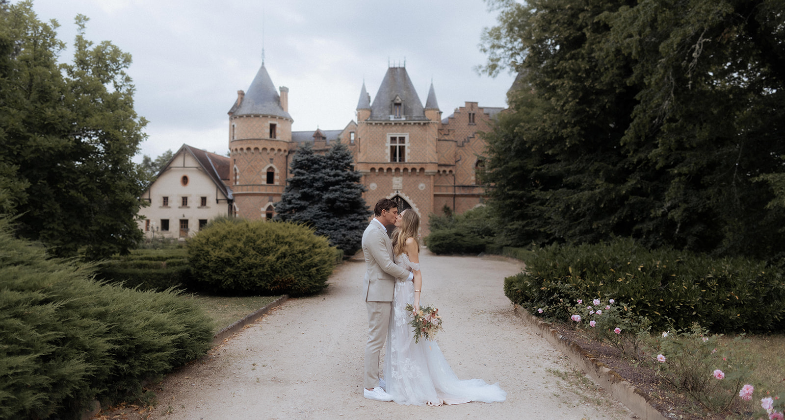Couple kisses on a gravel driveway before a pink-brick French chateau with turrets, bride holding terracotta-toned bouquet