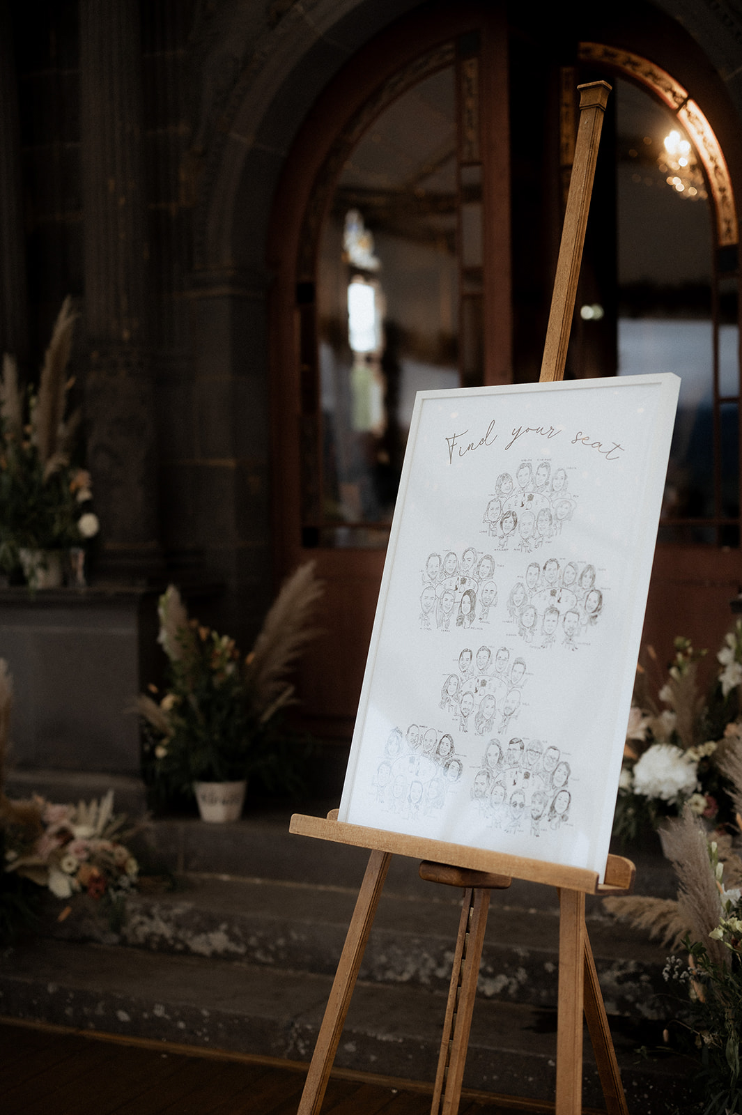 Illustrated seating chart on easel with pampas grass and white floral arrangements in stone-arched venue
