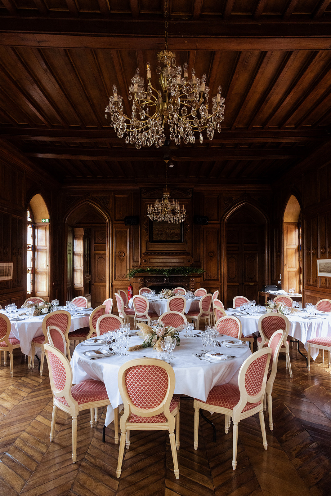 A wedding reception room set up for a seated dinner inside a château, shot as a wide interior portrait. The room features dark wood-paneled walls with Gothic-arched alcoves, a herringbone parquet floor, and two crystal and gilt chandeliers hanging from a coffered dark wood ceiling. Four round tables are dressed in white floor-length linens and set with crystal stemware, silverware, and folded napkins; centerpieces include dried pampas grass and low floral arrangements in muted cream and blush tones. The chairs are cream-painted Louis XVI-style medallion-back chairs upholstered in a terracotta-red diamond-pattern fabric. A carved stone fireplace dressed with greenery anchors the back wall. No guests are present. Potential venue feature image.