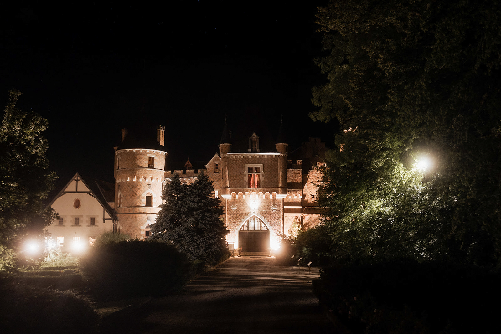 A wide-shot exterior photograph of a château-style castle venue taken at night, with no people visible. The building features a round turret tower on the left, a pointed Gothic arch entrance gate at the center, decorative brickwork facades, multiple chimneys, and smaller turret elements along the roofline. The structure is illuminated by warm amber uplighting that highlights the architectural details of the entrance and tower, while pathway lights guide a gravel driveway toward the main gate. A half-timbered outbuilding is visible to the left, also lit from below. The surrounding grounds are dark with large mature trees framing the composition on both sides. Potential venue feature image.