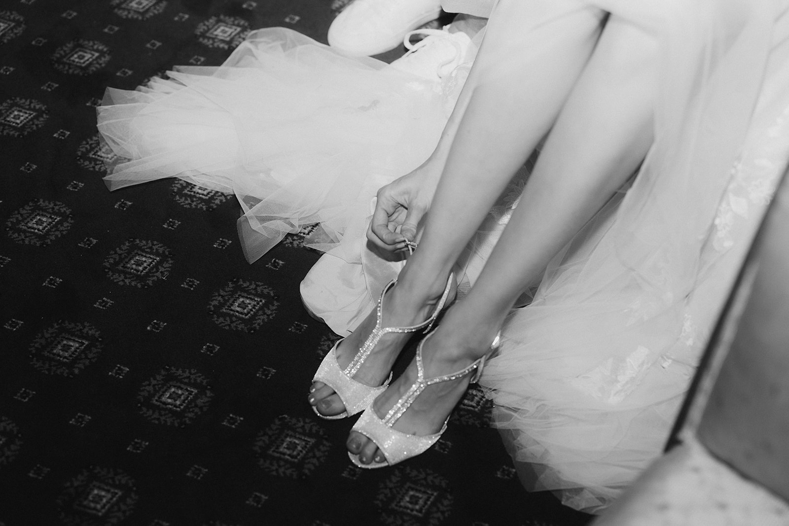 Black and white close-up of bride fastening crystal T-bar sandal strap with tulle skirt pooling on carpet