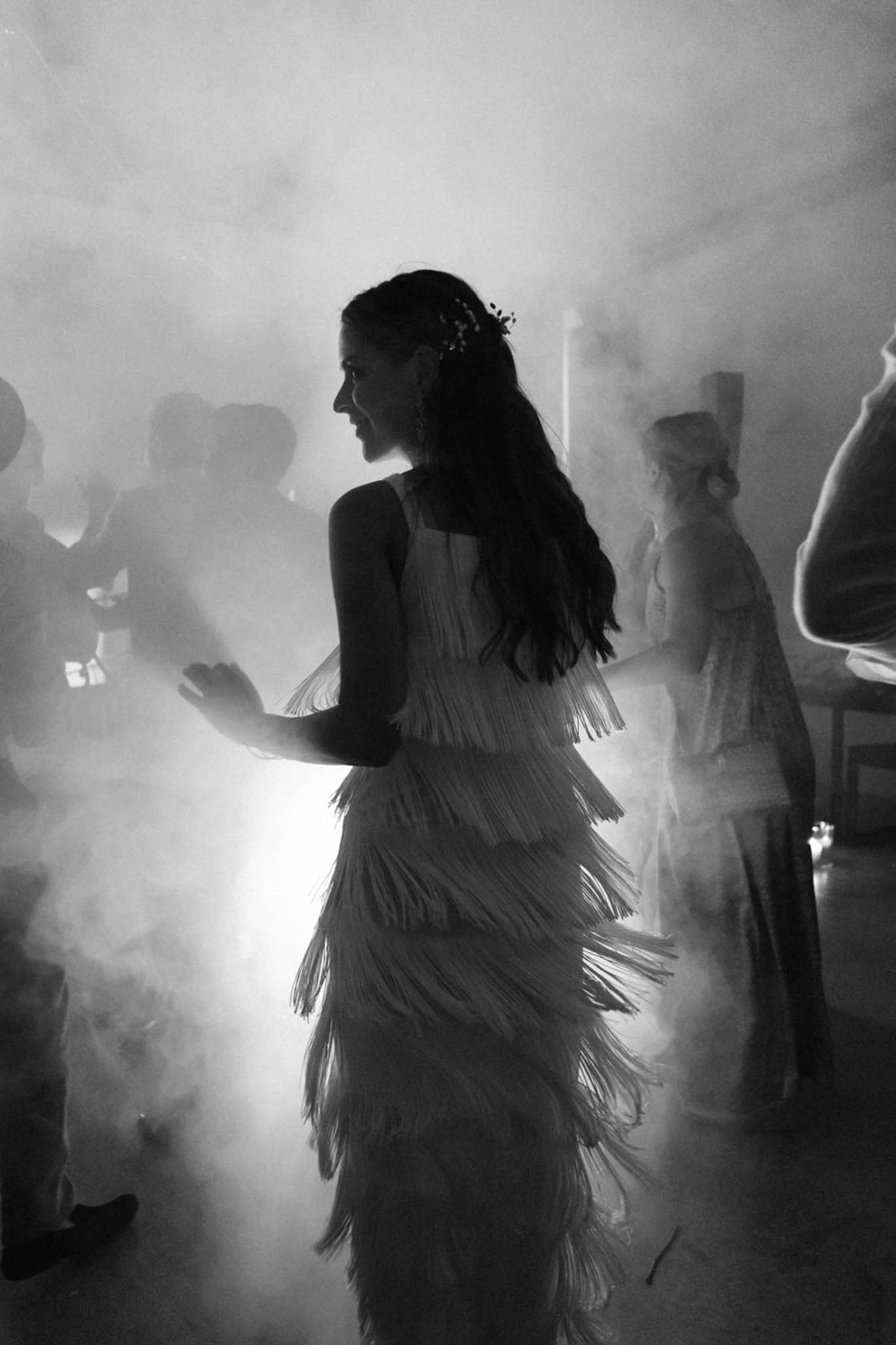 Black and white image of bride dancing in fringe dress surrounded by fog machine smoke with dramatic backlighting