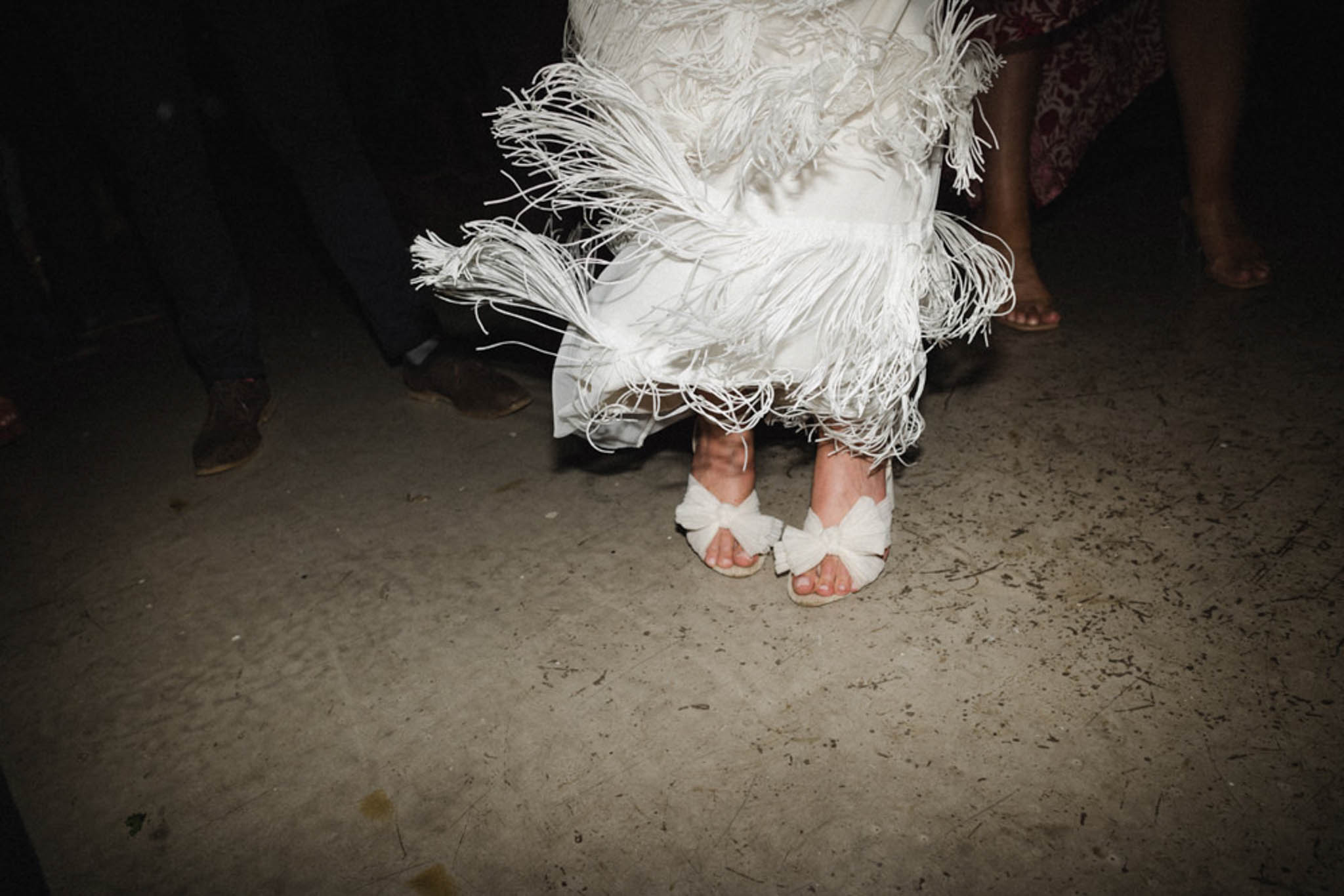 Close-up of bride's white bow sandals and fringed dress hem while dancing on a scuffed reception floor