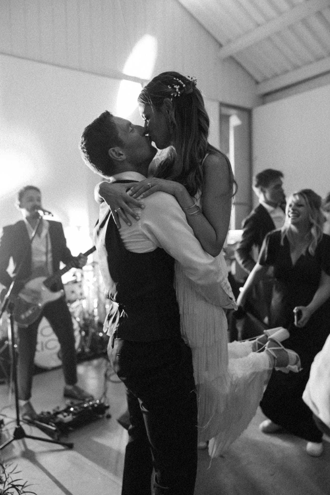 This black-and-white image captures the couple's first dance in an indoor reception space with a vaulted ceiling and white walls. The groom, wearing a dark waistcoat and trousers, lifts and kisses the bride, who wears a light-colored flowing dress with spaghetti straps and has her long hair half-up with small floral pins. A live band guitarist is visible performing on the left side of the frame, and approximately five to six guests can be seen in the background reacting with visible enthusiasm. The image has high contrast with soft ambient lighting, shot at mid-distance as a candid portrait-style frame capturing the energy of the reception.