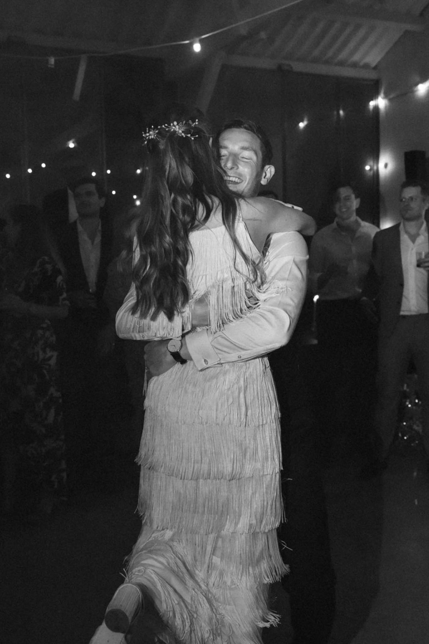 Black-and-white image of a couple sharing their first dance at an indoor evening reception, likely in a barn or covered venue with an exposed roof structure overhead. The bride wears a tiered fringe dress with long sleeves and has long wavy hair adorned with a delicate floral hair piece, while the groom, laughing with eyes closed, wears a white jacket with snap buttons and a watch. The pair embrace closely on the dance floor as several guests watch and smile in the background. Festoon/bistro lights are strung across the ceiling, creating warm bright points of light against the dark interior, giving the scene a boho-festive atmosphere.