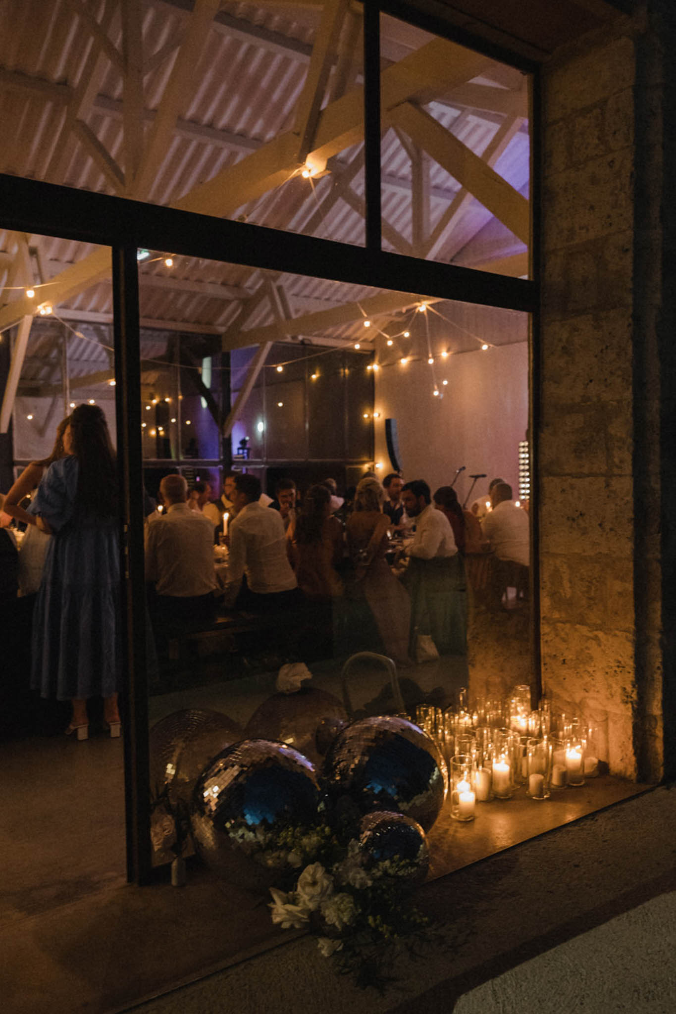 Evening reception through glass doors of converted barn with disco balls, candles, and festoon lights