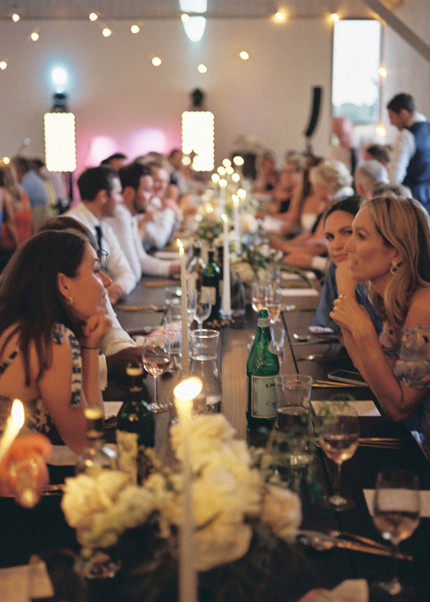 Guests seated at long banquet table during reception speeches in a barn venue with fairy lights and candlelight