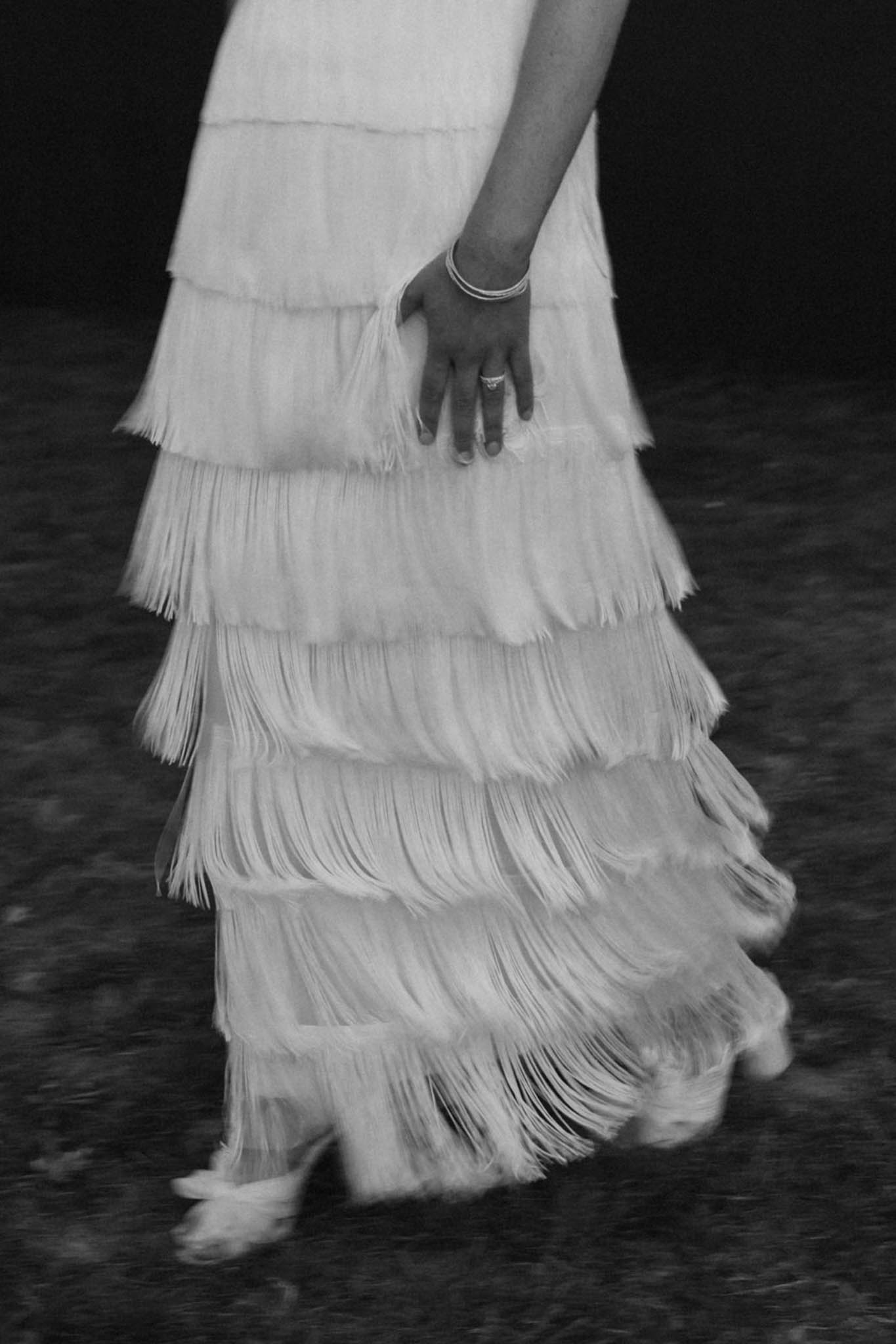 This black-and-white detail shot captures the lower half of a bride in motion, showcasing a floor-length wedding dress featuring multiple tiers of long fringe that cascade from hip to hem. The movement of the fringe creates a slight blur, emphasizing the dress's texture and flow against a dark background. The bride's hand is visible at her side, adorned with a diamond engagement ring and several thin stacking bracelets. The image is a close-up portrait-style crop focused entirely on the dress design and accessories, with high contrast between the light fringe tiers and the dark surroundings.