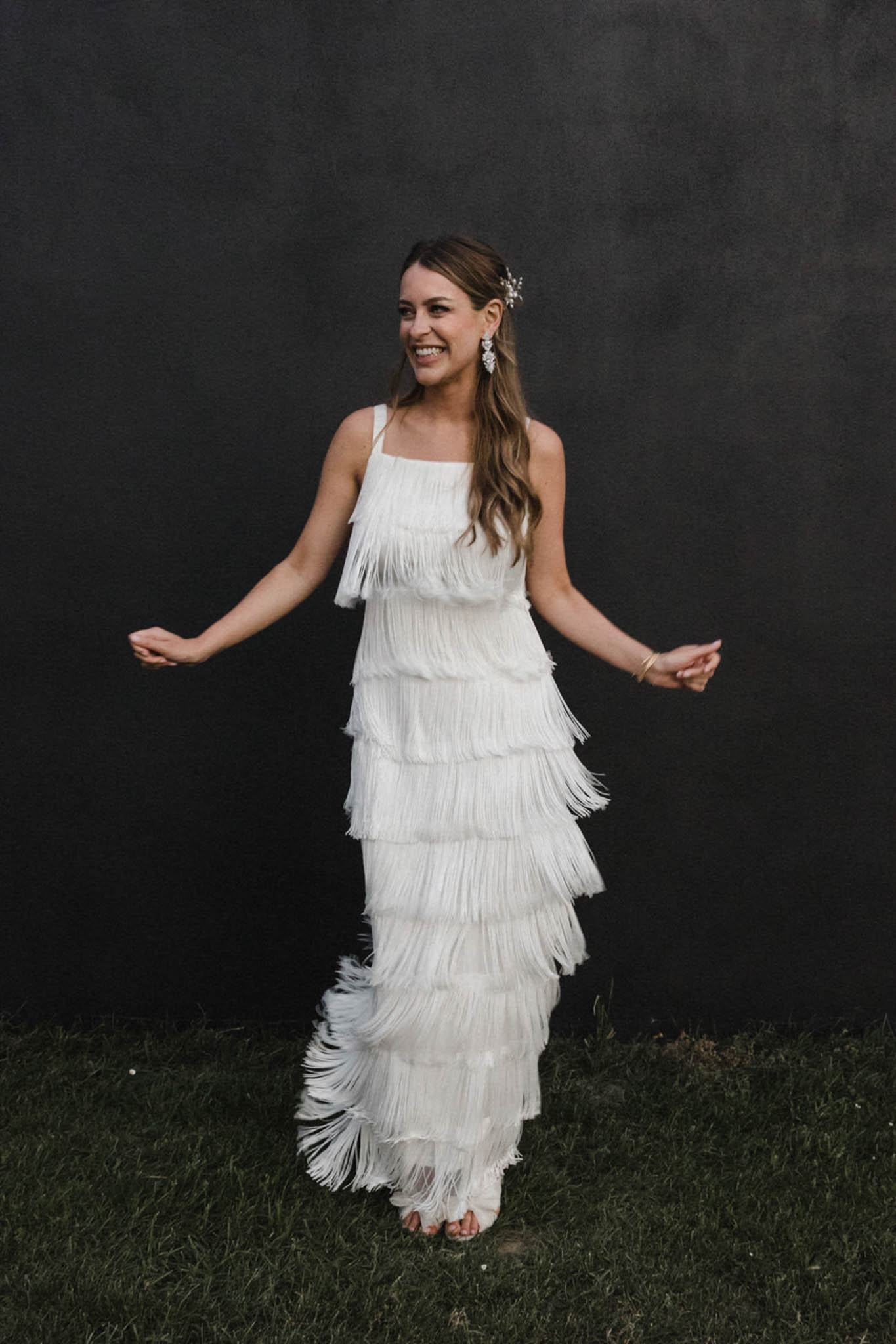A bride poses alone outdoors against a dark charcoal wall, laughing and holding her arms slightly out to her sides. She wears a floor-length white tiered fringe wedding dress with thin spaghetti straps and a square neckline, featuring multiple horizontal layers of fringe from bodice to hem in a 1920s-inspired flapper style. Her accessories include large crystal drop earrings, a delicate crystal hair pin worn in her loose wavy brown hair, and a gold bangle bracelet. She has open-toe heeled sandals visible at the hem. The shot is a full-length portrait with a dark, almost black background that contrasts sharply with the white dress, giving the image a high-contrast, editorial feel.