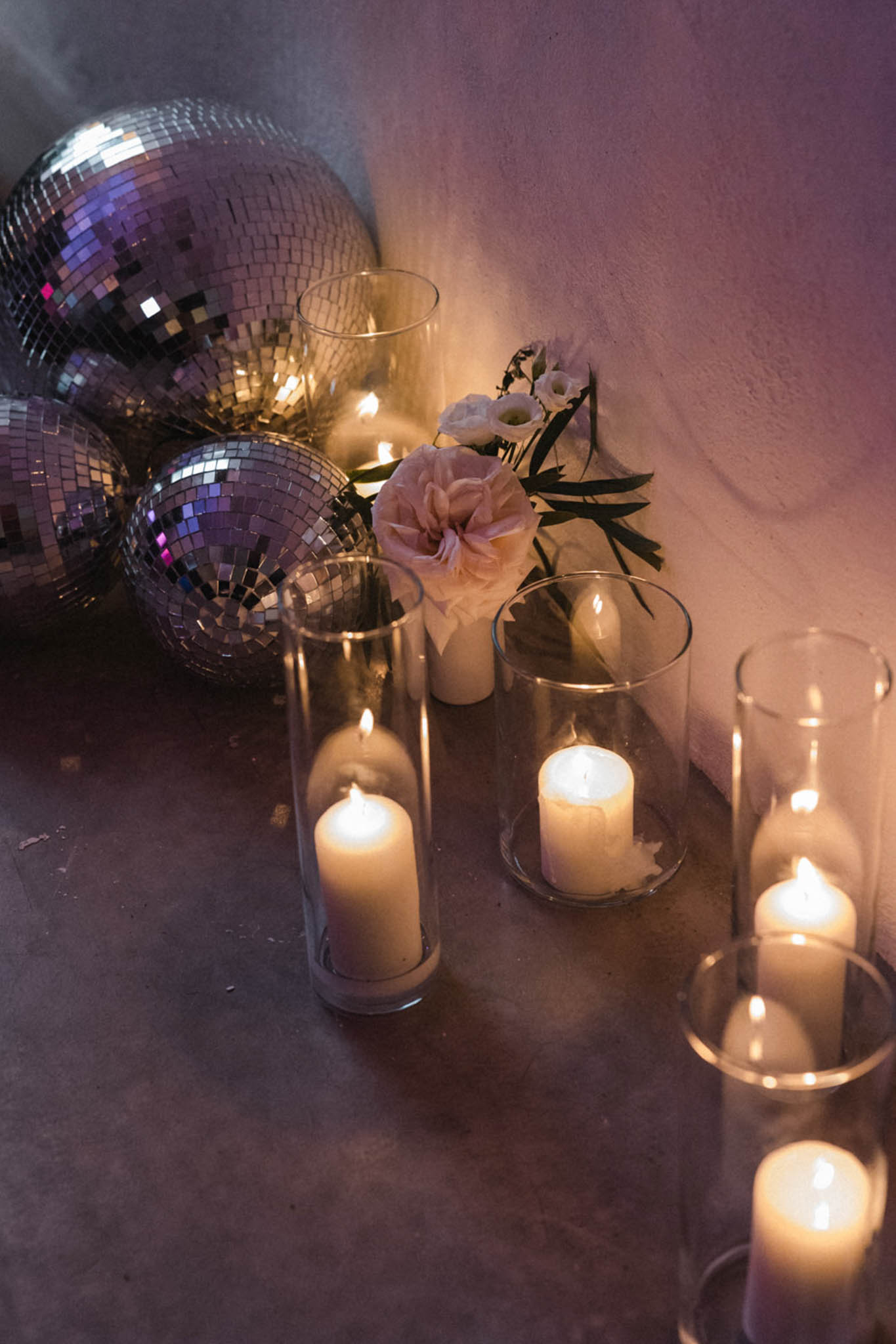Pillar candles in glass hurricanes beside silver disco balls and blush garden rose arrangement on floor