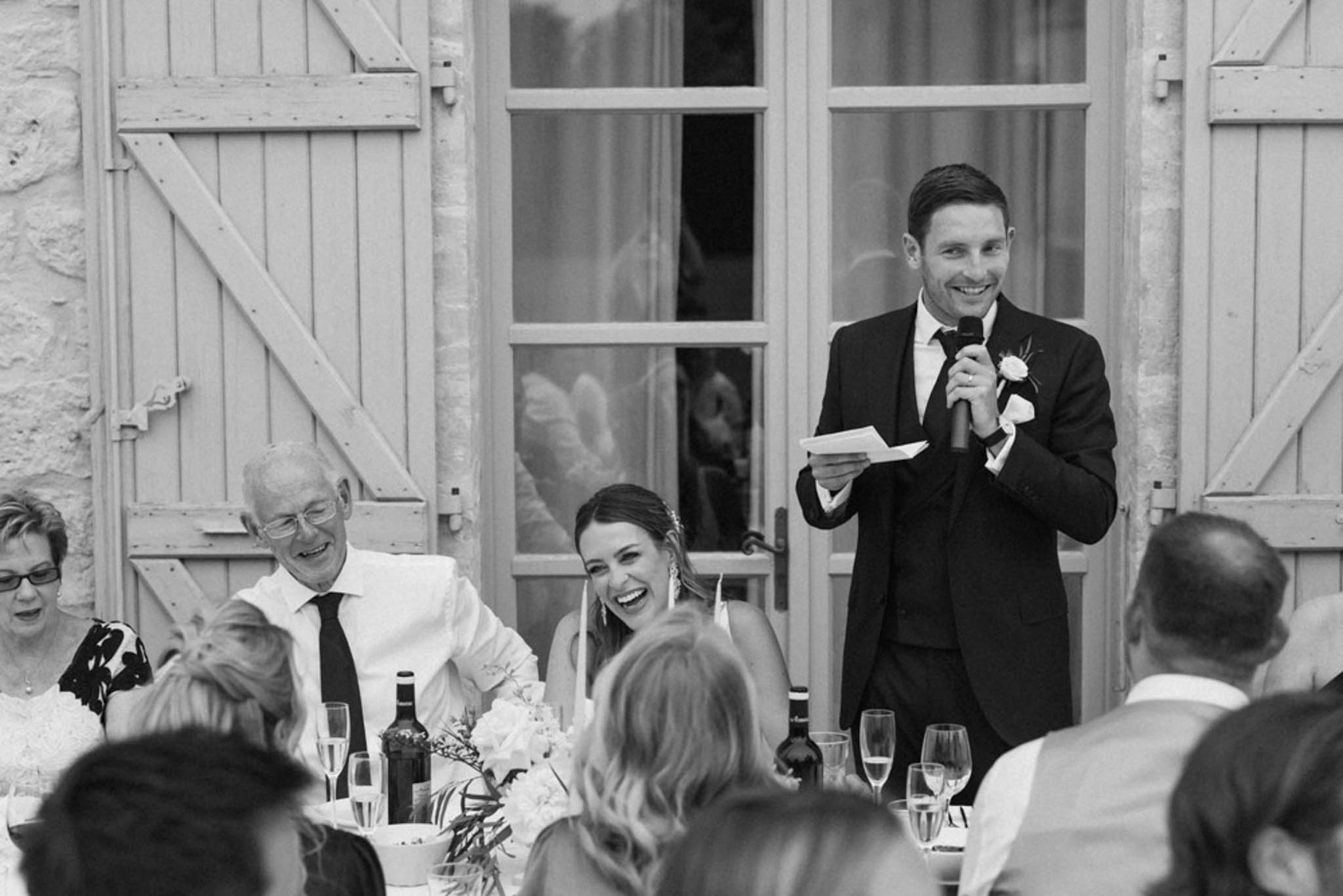 Black and white candid of wedding speech with bride laughing beside guests at outdoor chateau reception