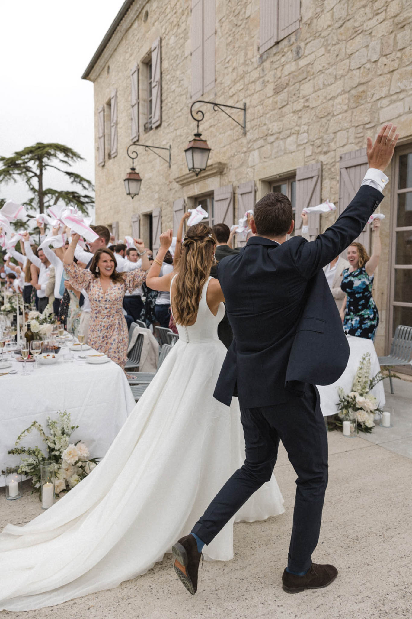 Bride and groom enter reception as guests wave white napkins at long tables before limestone building