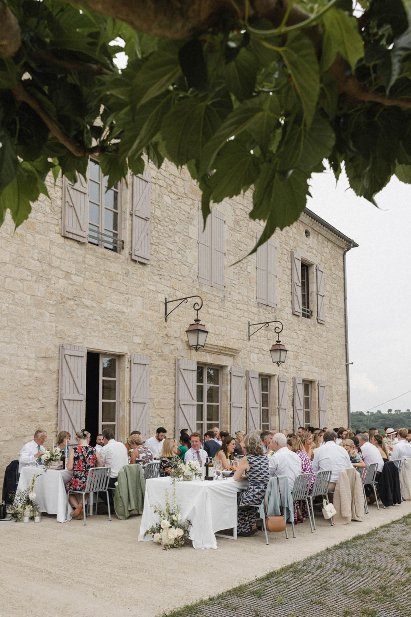 An outdoor wedding reception dinner is underway in front of a two-storey French stone manor house with taupe-grey shutters and wrought-iron lanterns mounted on the facade. Approximately 60–80 guests are seated along long banquet-style tables covered in white linen, arranged in rows directly against the building on a gravel terrace. Table centerpieces feature white florals — likely roses or ranunculus — with green foliage in low arrangements, and wine bottles and glassware are visible on the tables. Guests are dressed in smart-casual summer attire including floral dresses, suits, and light linen, and the overall styling is relaxed and classic with a neutral and white decor palette. The wide shot is framed from a slight distance with fig tree branches in the foreground, capturing the full scene and building facade. Potential venue feature image.