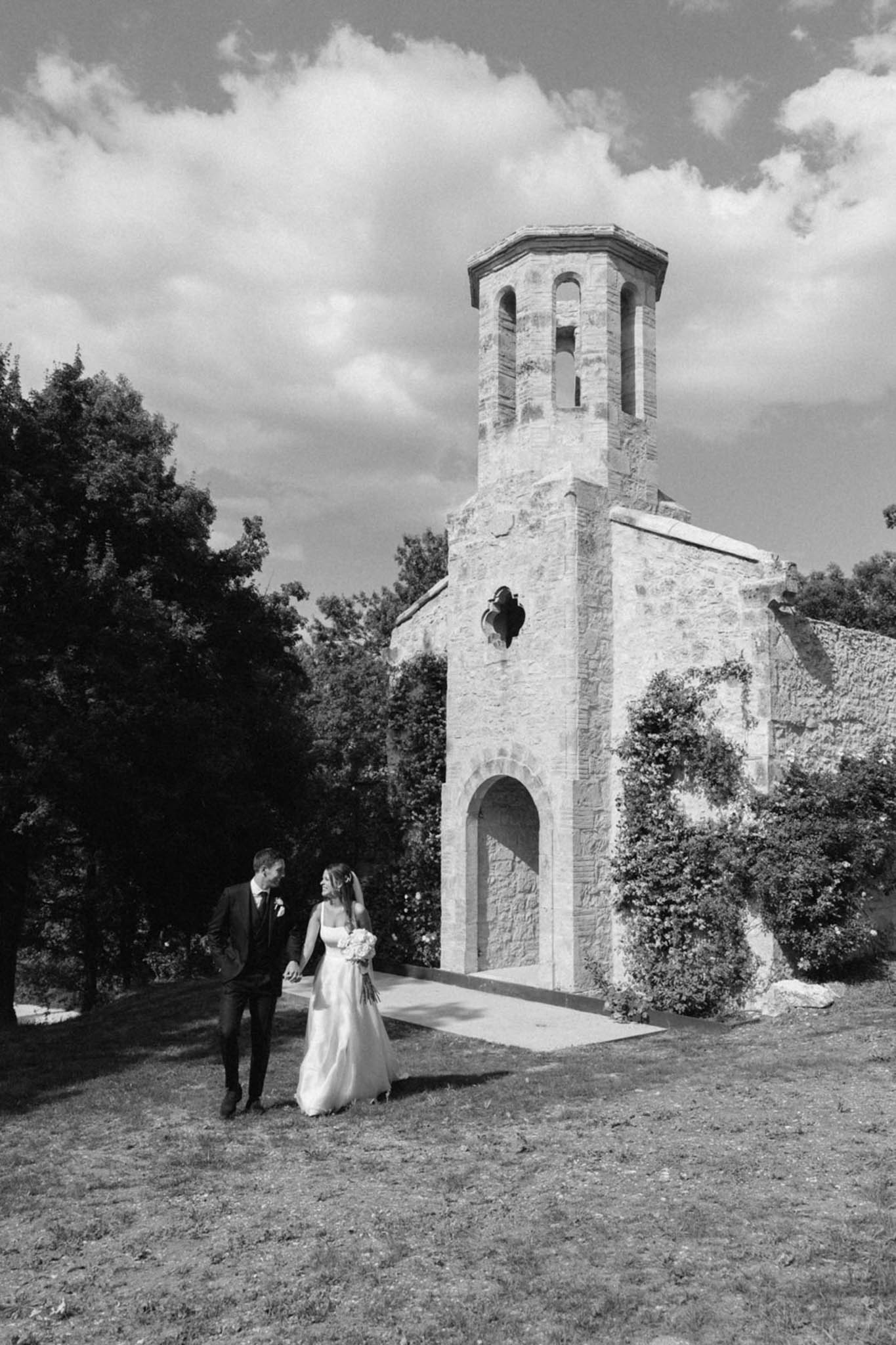 Bride and groom walking hand-in-hand before Romanesque stone chapel with bell tower in B&W