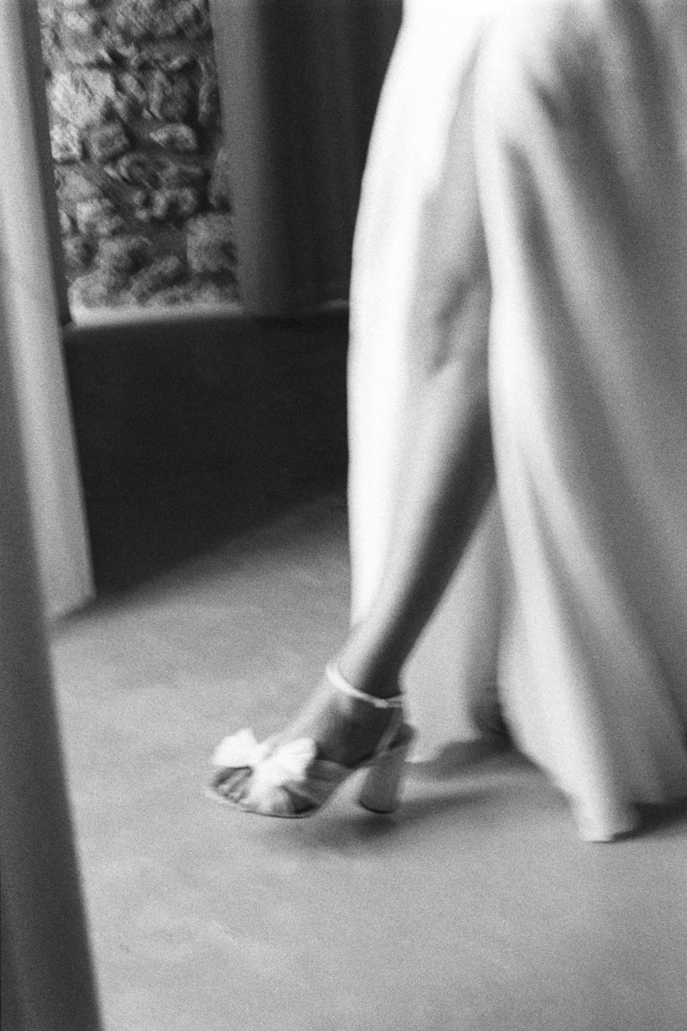 A black-and-white close-up detail shot focusing on the lower half of a bride's figure as she walks or moves indoors. The image captures a strappy block-heel sandal with a knotted toe detail, partially visible beneath the flowing skirt of a full-length wedding dress. The composition shows soft mid-tones and gentle contrast, with the fabric of the dress slightly motion-blurred, suggesting movement. A stone wall is partially visible in the background, indicating an interior space with historic architectural features.