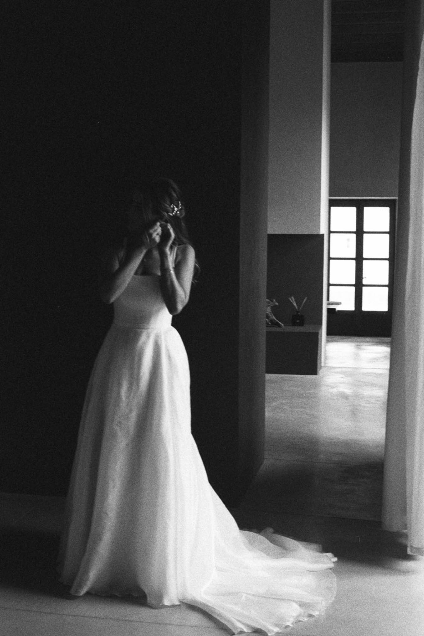 Black and white image of bride adjusting hair accessory in minimalist room with dramatic window light and trailing gown