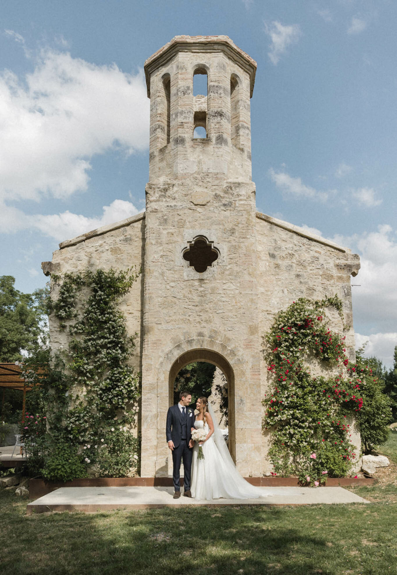 A couple portrait taken outdoors in front of a small Romanesque stone chapel featuring an octagonal bell tower, a quatrefoil window, and an arched entrance doorway. The groom wears a navy three-piece suit with a tie and boutonnière, while the bride wears a strapless ivory ball gown with a full tulle skirt and carries a blush and white bouquet. The two stand facing each other on a stone platform beneath the chapel's arched entrance, flanked by climbing roses in deep red and white on either side of the facade. The composition is a full wide shot that centers the chapel architecture prominently with the couple positioned in the lower third. Potential venue feature image.