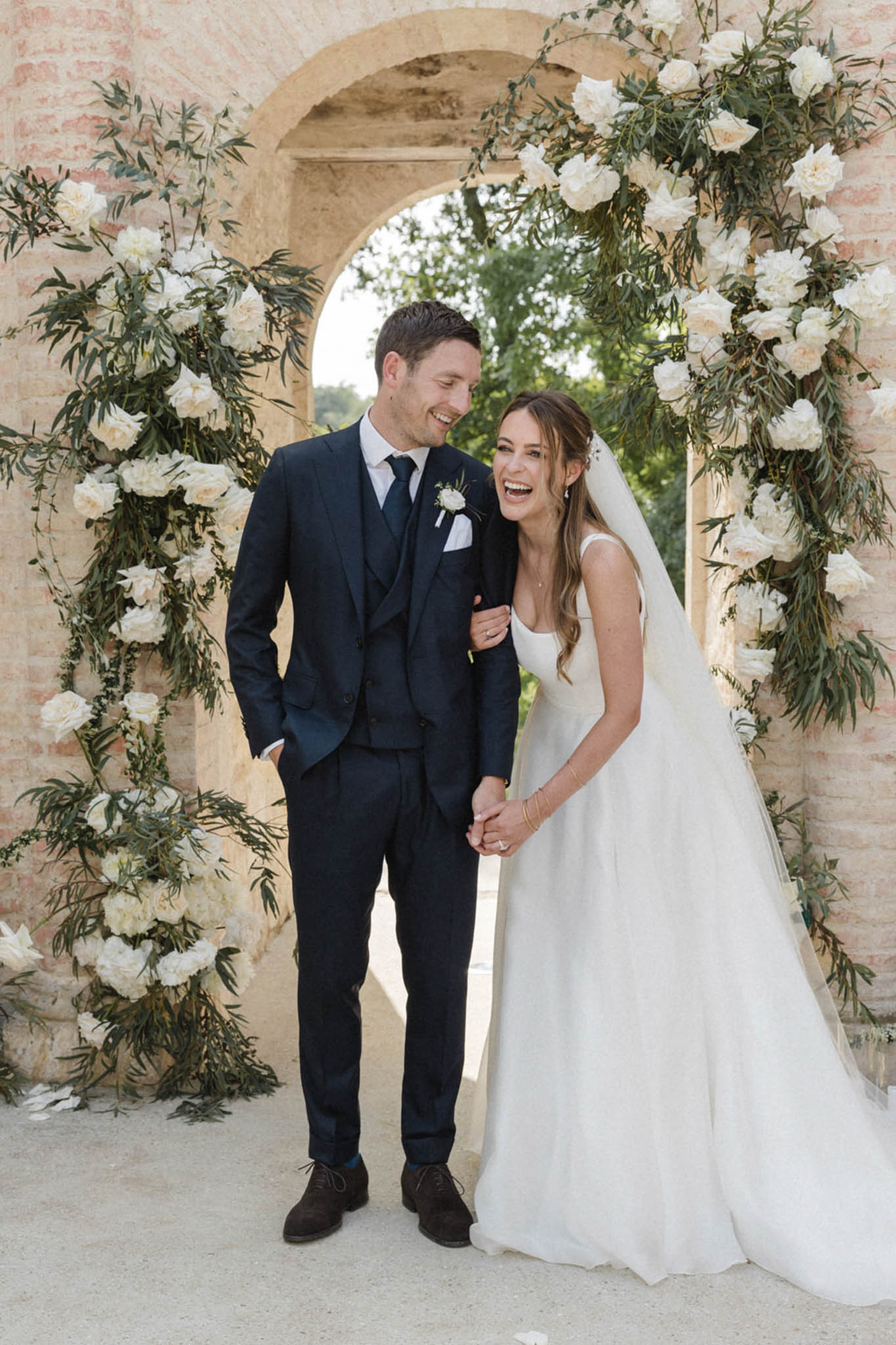 Laughing bride and groom under stone archway decorated with ivory roses and trailing olive branches