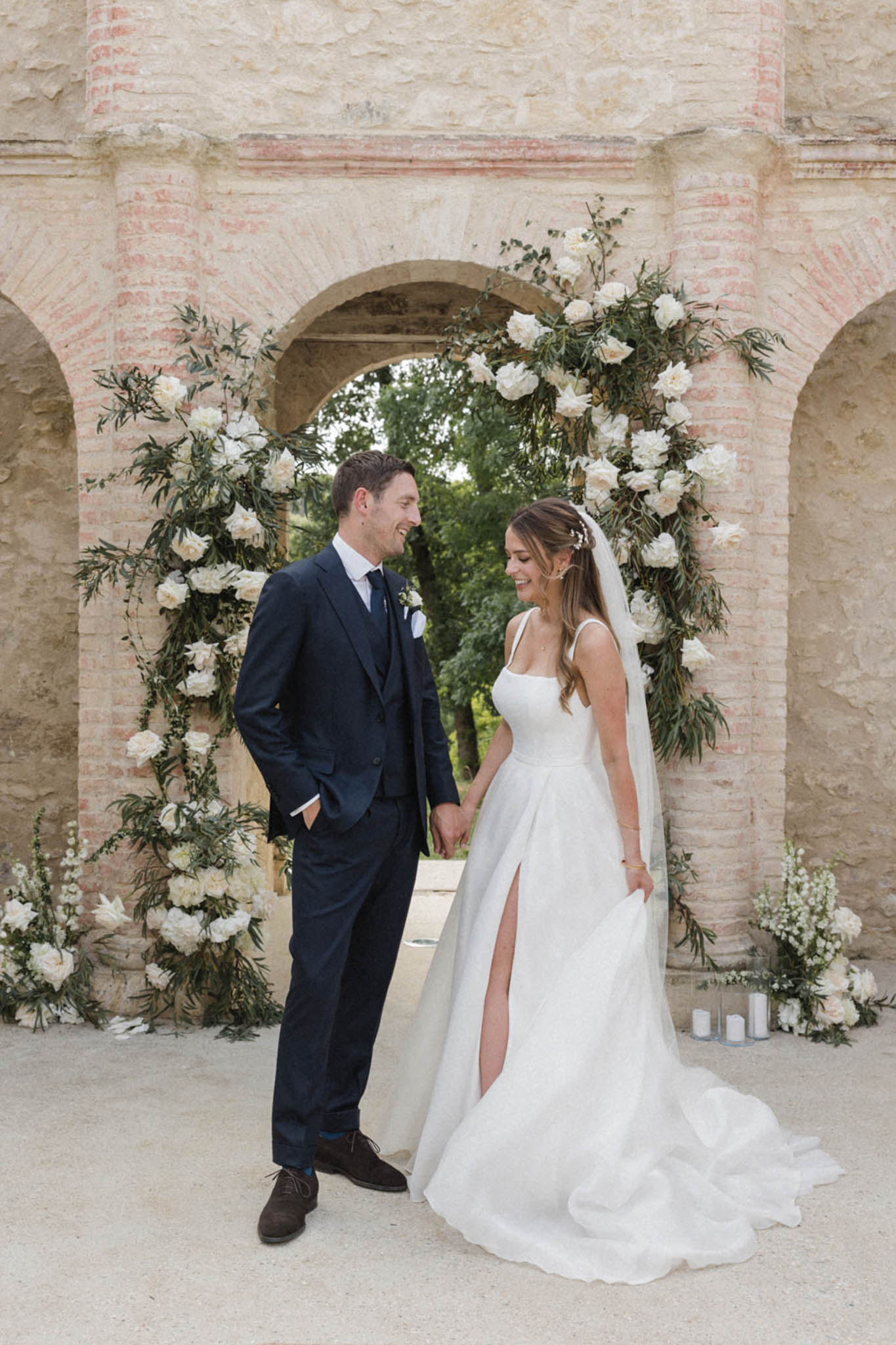 A couple portrait taken outdoors in front of a stone and brick archway structure, likely at a French château or historic estate. The couple holds hands and laughs together, with the groom in a navy three-piece suit with a navy tie and a white boutonnière, and the bride in a white A-line gown with thin square-neck straps, a front slit, and a cathedral-length veil with a floral hair accessory. Behind them, a large circular floral arch is adorned with white peonies, white roses, and trailing greenery including olive-style branches, framing the stone arch opening. Small glass cylinder candles and additional white floral arrangements are placed at the base of the arch on either side. The styling is modern classic with a white and green floral palette. Medium full-length portrait shot.