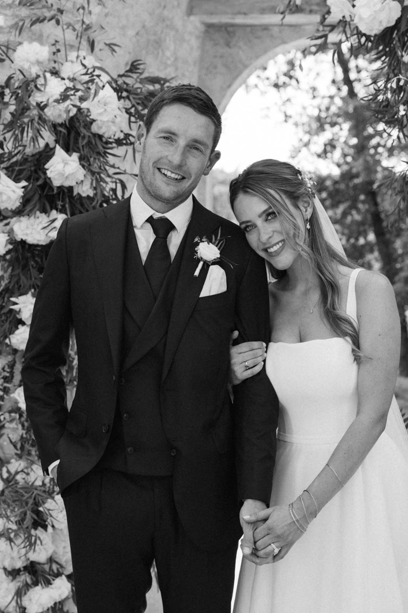 A black-and-white couple portrait taken outdoors, showing the bride and groom posing together in front of a large floral arch and a stone archway. The groom wears a dark three-piece suit with a dark tie, white pocket square, and a rose boutonnière; the bride wears a simple sleeveless gown with a square neckline, a cathedral-length veil, floral hair accessories, drop earrings, and stacked bracelets. Both are smiling directly at the camera, with the bride leaning her head against the groom's shoulder and holding his hand. The floral arch behind them features large full blooms — likely peonies or garden roses — with trailing foliage, rendered in bright whites and soft mid-tones against the high-contrast B&W processing. The composition is a mid-length portrait with soft background blur.