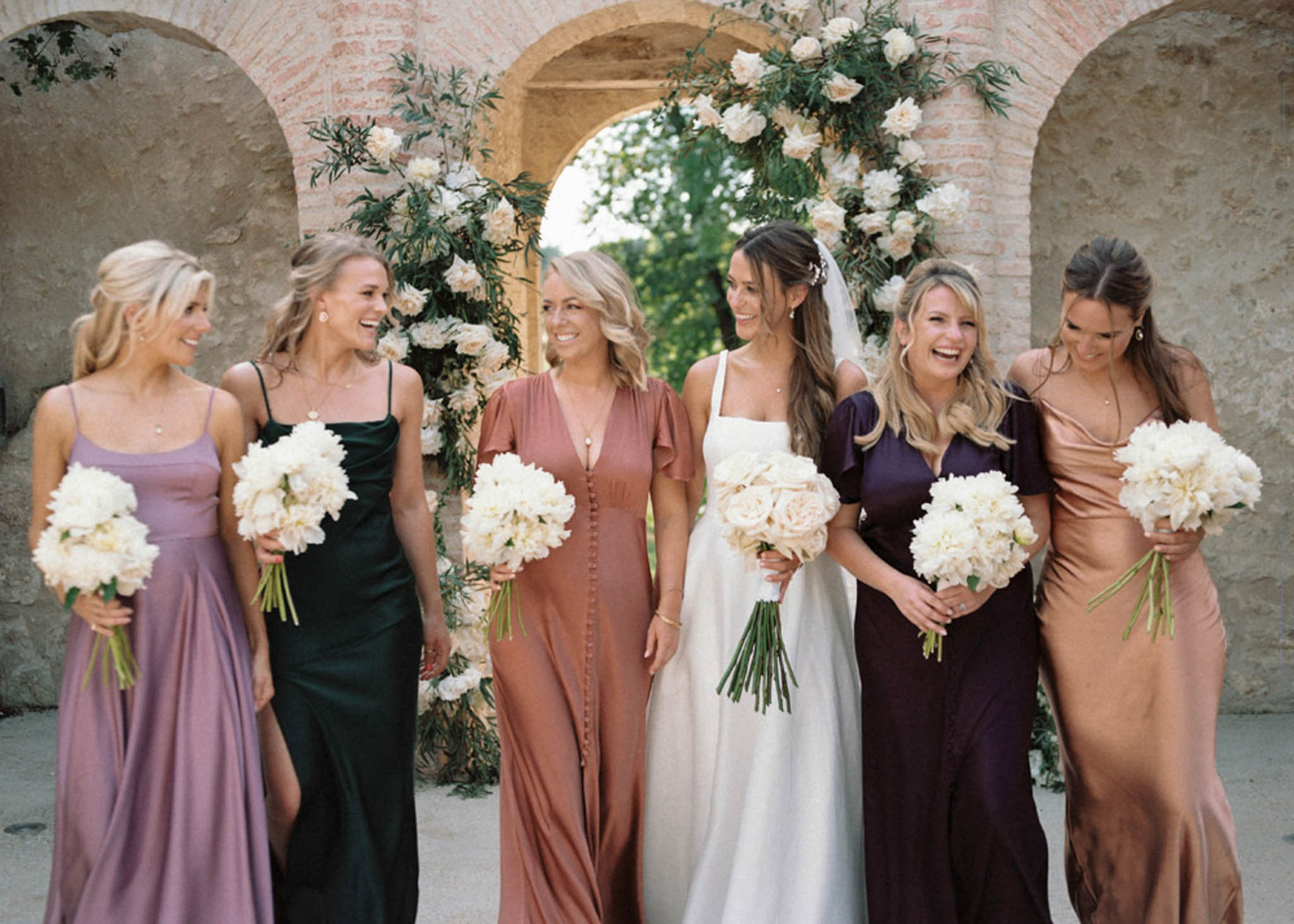 Bride with five bridesmaids in mismatched jewel-tone dresses before circular floral arch at stone arcade