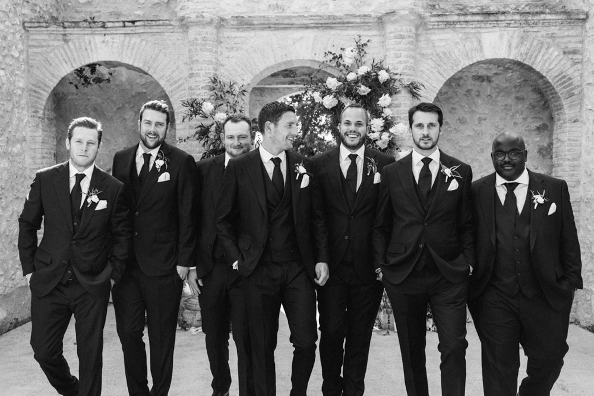 Black and white groom and six groomsmen in dark suits laughing before triple-arched stone wall with peonies