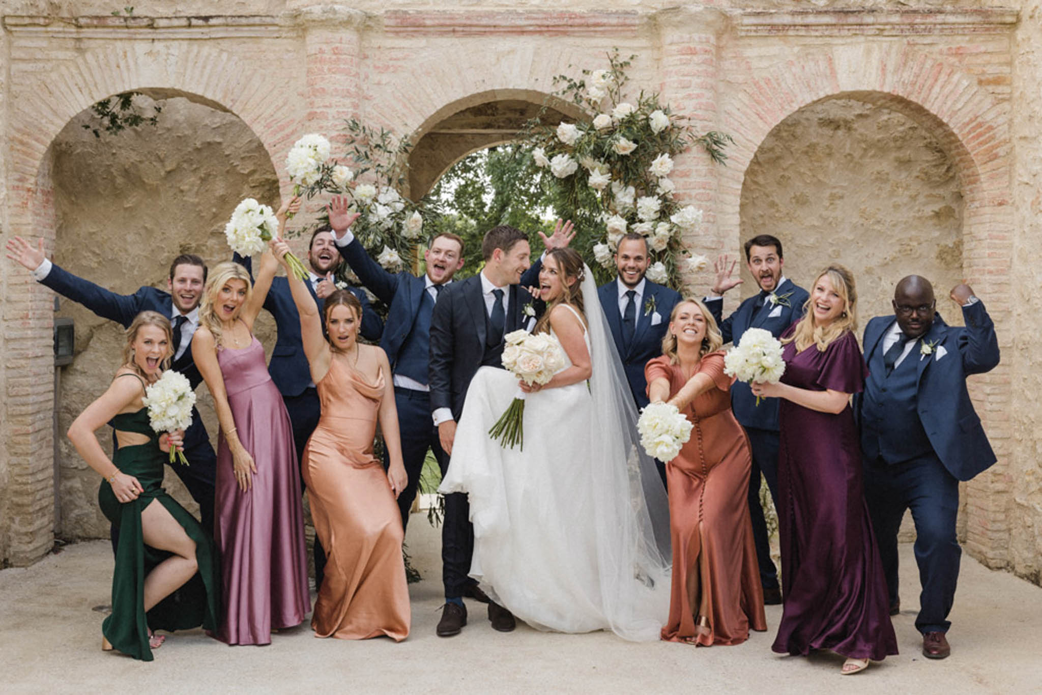 Thirteen-person bridal party celebrates under stone arcade with white peony arch and mismatched satin gowns