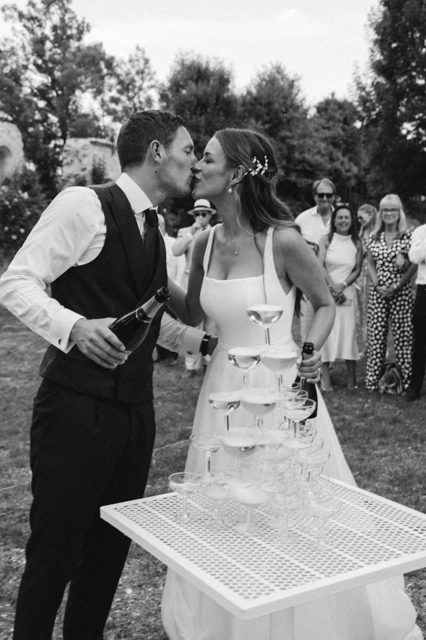 Black and white photo of bride and groom kissing beside champagne tower during outdoor cocktail hour
