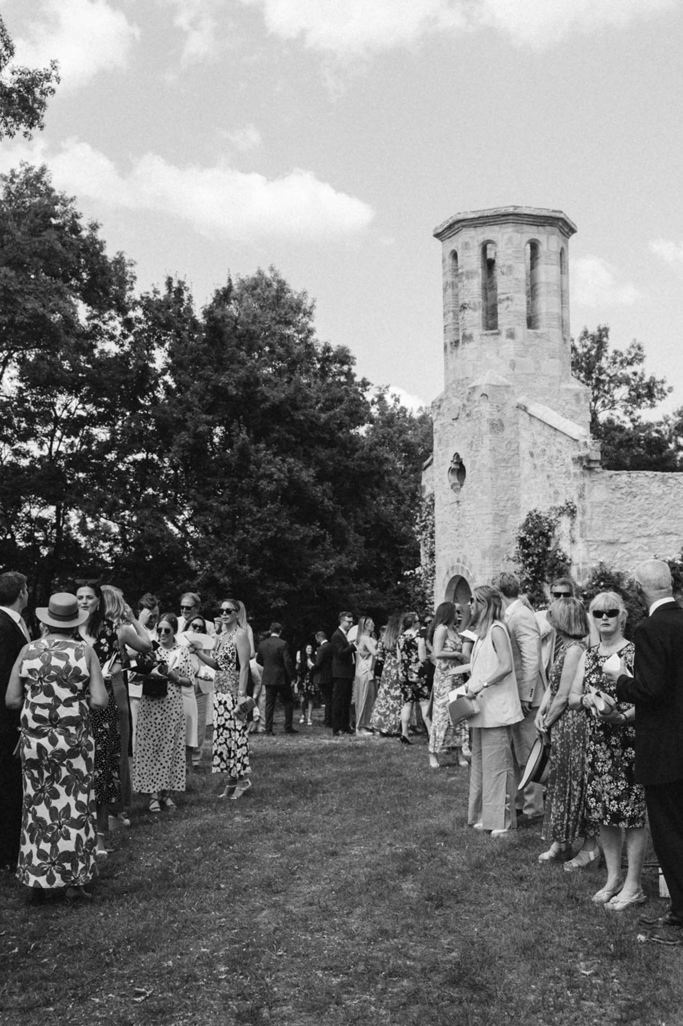 Wedding guests mingling on lawn before Romanesque stone chapel with octagonal bell tower in black and white