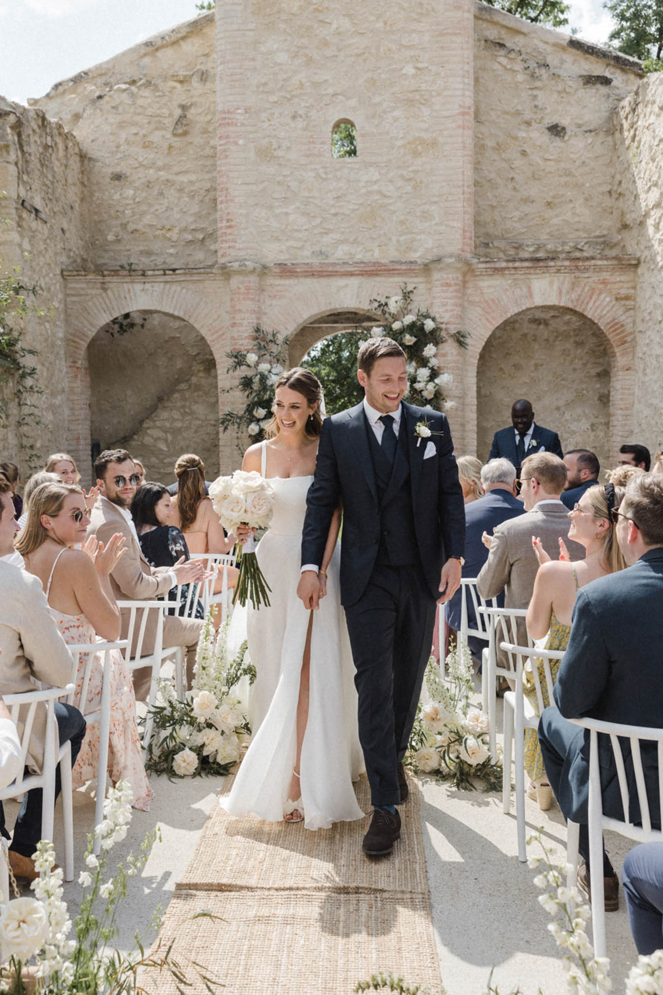 The bride and groom walk back down the aisle together following their outdoor ceremony, both smiling, in a wide portrait-style shot. The ceremony takes place within the open-air ruins of a stone chapel or abbey, with exposed brick arched openings and weathered limestone walls forming the backdrop. The bride wears a sleek ivory slip-style gown with a front slit and thin straps, carrying a bouquet of white garden roses and greenery with long trailing stems. The groom wears a navy three-piece suit with a white pocket square and a white floral boutonnière. Aisle decor consists of low clusters of ivory roses, white delphiniums, and greenery lining a natural jute runner, with a floral arch of white roses and foliage framing the central arched opening behind them. Guests — approximately 40–50 visible — are seated on white chairs on either side of the aisle, many applauding. The overall decor palette is ivory and white with natural greenery, consistent with a classic, clean aesthetic. Potential venue feature image.
