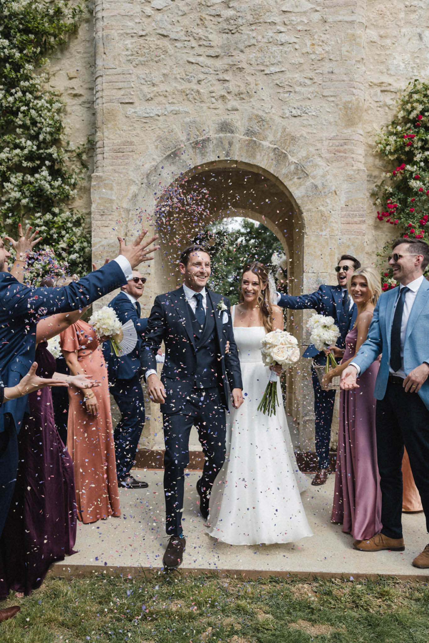 Bride and groom walk through confetti exit under stone archway with guests throwing colourful paper confetti