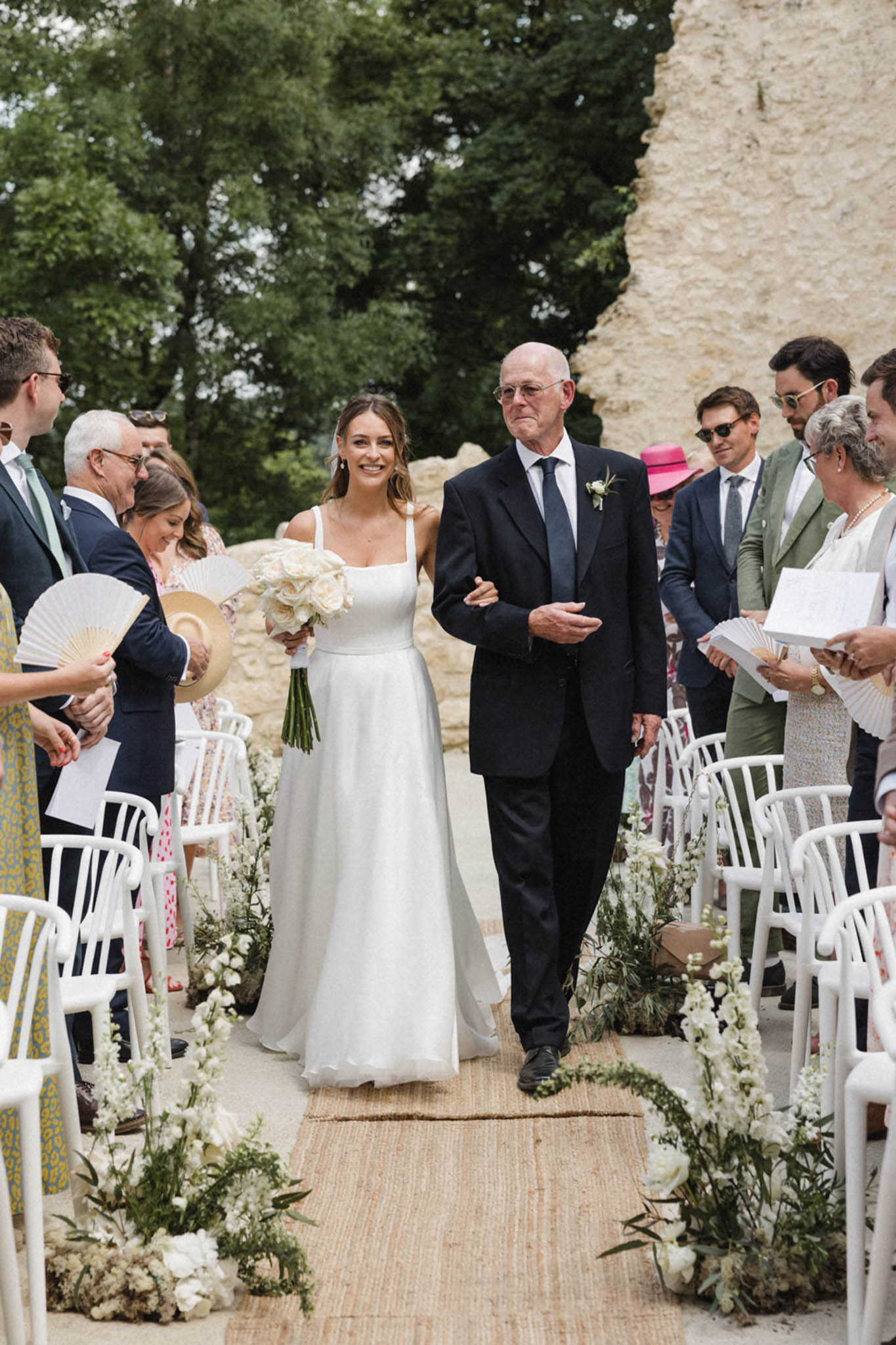 The bride walks down the aisle escorted by an older man, likely her father, during an outdoor ceremony. The bride wears a simple A-line white gown with square neckline and thin straps, carrying a bouquet of ivory roses with greenery stems. The aisle is lined with a natural jute runner and low floral arrangements of white delphiniums, white roses, and trailing greenery placed directly on the ground, with a small floral arch detail at the base. White bentwood chairs seat approximately 30–40 guests on either side, several of whom are holding white paper hand fans and straw hats, suggesting warm weather. The setting is an outdoor ceremony space adjacent to a limestone or tuffeau stone structure, styled with a clean, natural aesthetic. The shot is a medium-wide portrait taken from ground level looking up the aisle toward the couple.