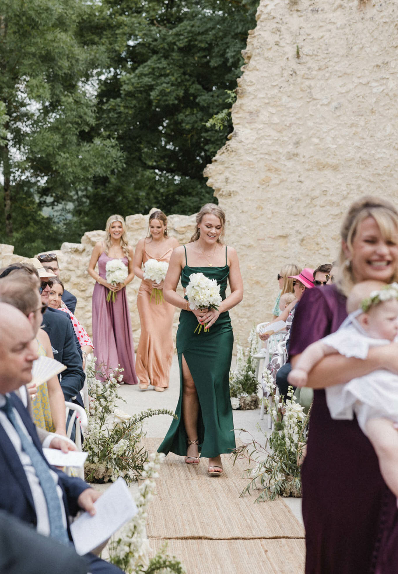 Three bridesmaids walk down the aisle during an outdoor wedding ceremony set against a ruined stone wall. The bridesmaids wear mismatched satin slip dresses in forest green, copper/champagne, and dusty mauve, each carrying a bouquet of white peonies with green stems. The aisle is lined with low white floral arrangements including white stock and trailing greenery laid along a natural jute runner. Seated guests on both sides watch the procession, including a woman in a pink visor on the right holding an infant. The shot is taken from a mid-distance, slightly elevated angle looking down the aisle toward the bridesmaids.