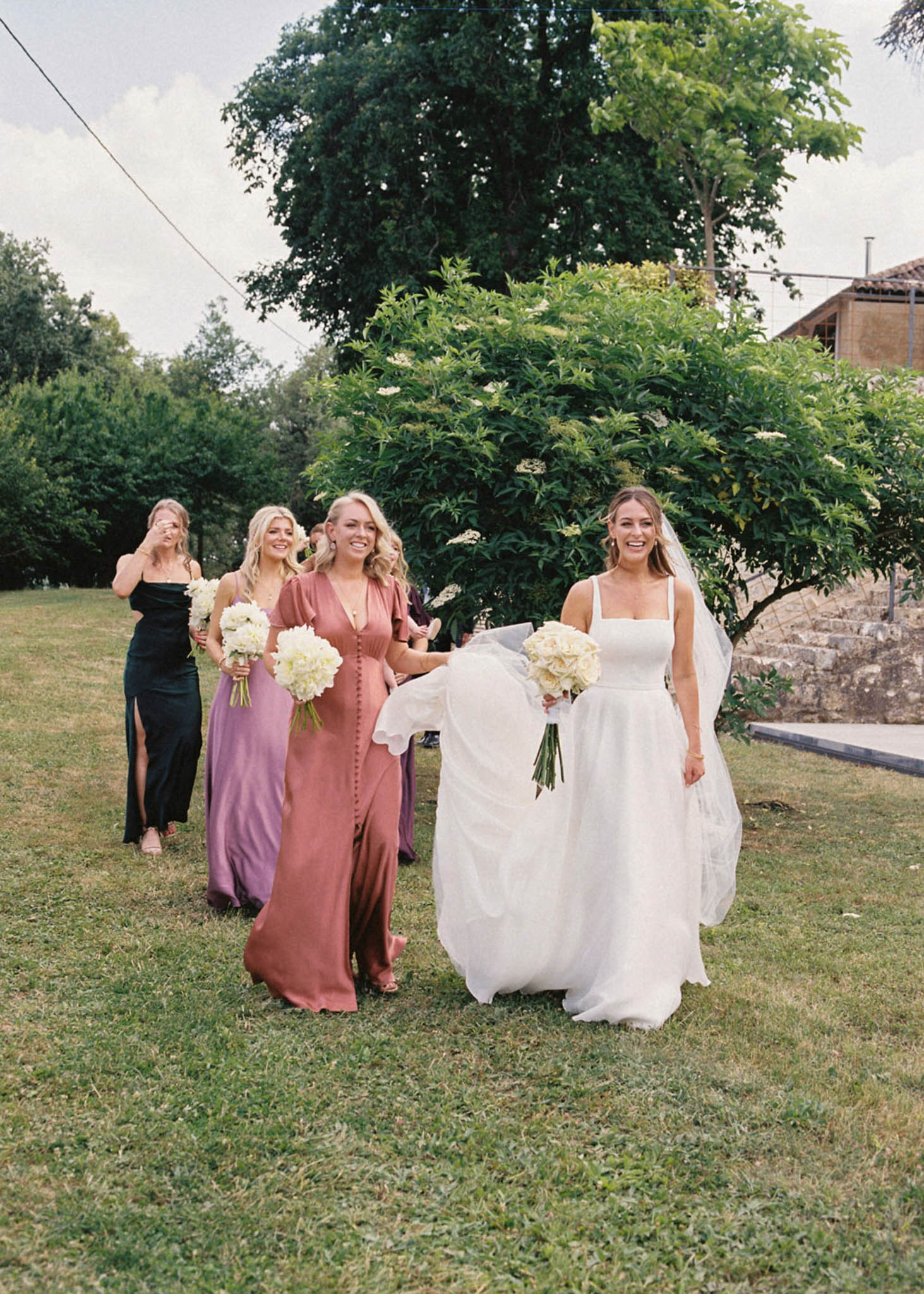 Bride and four bridesmaids in mismatched dusty rose, mauve, green, and lilac dresses walking on lawn