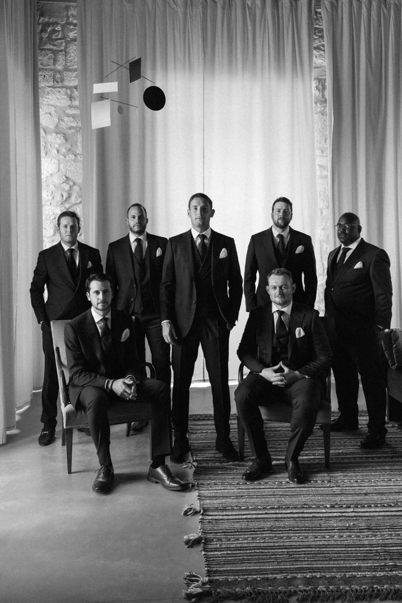 Black and white group portrait of groom and six groomsmen in dark suits posed indoors with hanging mobile sculpture