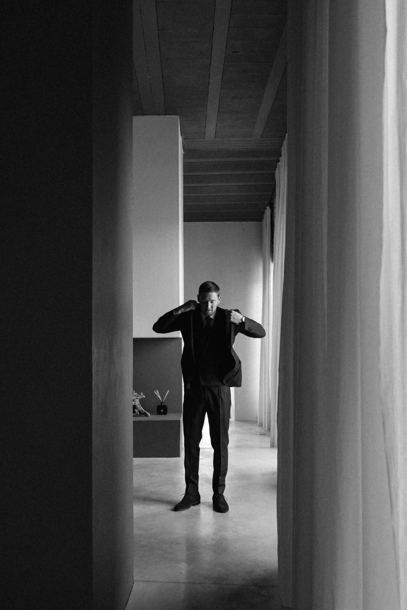 Black-and-white getting-ready portrait of a groom adjusting the lapels of his dark suit jacket while standing at the end of a narrow interior corridor. The image is shot in high contrast with deep shadows framing the foreground and the groom backlit by light from the room beyond, creating a strong silhouette effect. The setting is a modern minimalist interior with smooth rendered walls, a wood-beamed ceiling, polished concrete floors, and sheer white curtains to the right; a low built-in shelf with a reed diffuser and small decorative object is visible to the left of the figure. The composition is a full-length portrait taken from a distance, using the corridor walls as natural framing to draw the eye toward the groom.